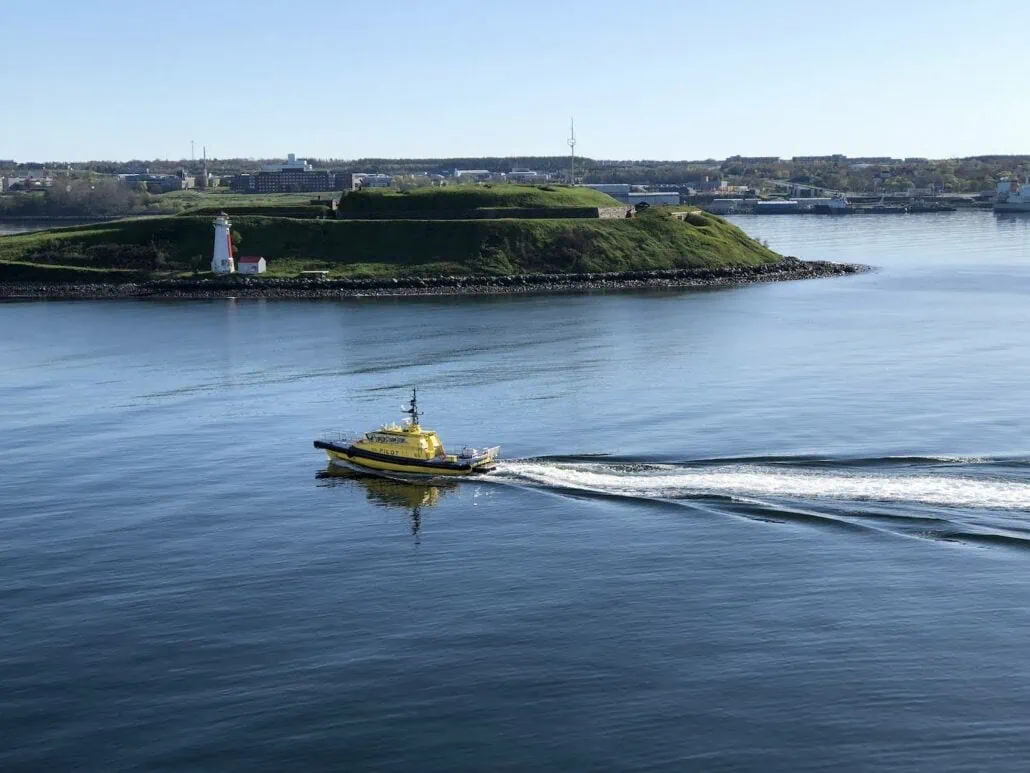 A yellow tugboat glides through the calm blue waters near Halifax Cruise Port, leaving a white trail in its wake. In the background, a small green island with a white lighthouse and buildings is visible under the clear sky—perfect for those seeking what to do in this picturesque setting.