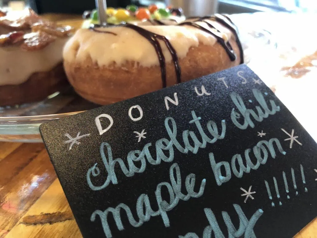 Close-up of a tray with two doughnuts at Halifax Cruise Port. A handwritten sign in front reads "DONUTS Chocolate Chili Maple Bacon" with decorative stars and exclamation marks. The doughnuts, perfect for those enjoying docked activities, are topped with frosting, chocolate drizzle, and colorful candies.