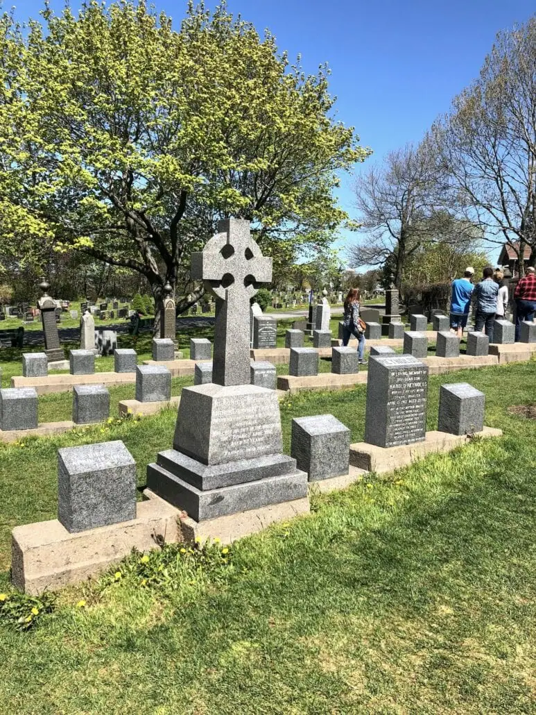 A cemetery scene with a row of gray tombstones, including a prominent Celtic cross, lies peacefully under the clear blue sky. Nearby, lush trees stand with fresh leaves. In the distance, a group of people from the docked Halifax Cruise Port walks by, soaking in the serene atmosphere.