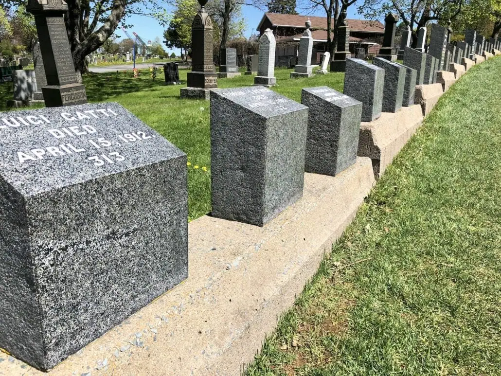 A row of gray stone grave markers, reminiscent of those near the Halifax Cruise Port, stand aligned on a concrete base, extending into the distance. The headstone in the foreground has an inscription dated April 15, 1912. Green grass and trees surround this solemn scene.