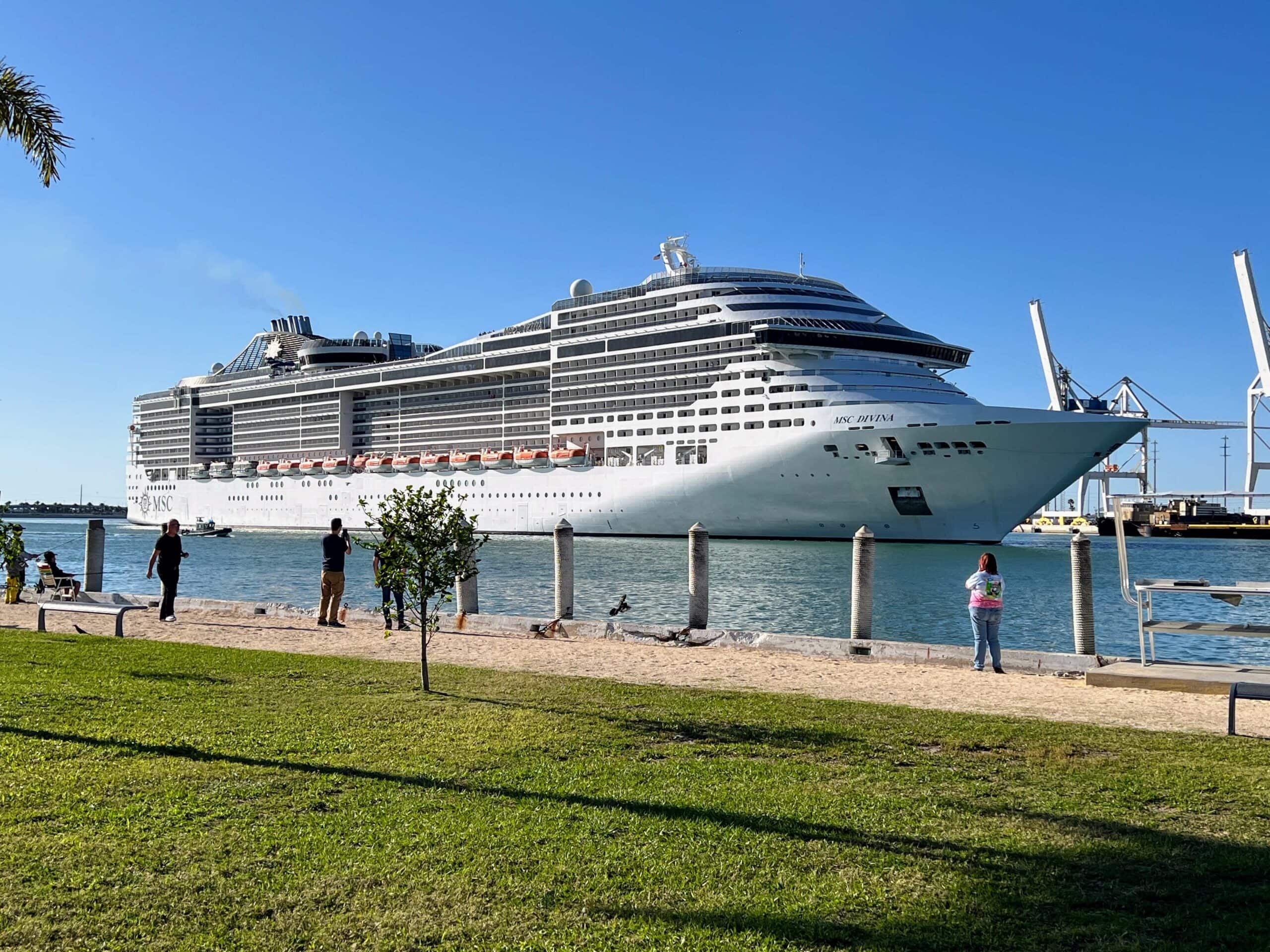 A large cruise ship is docked near a shoreline with a grassy area and several people standing nearby, all under a clear blue sky. Towering industrial cranes rise in the background, indicating the busy activity at Port Canaveral.