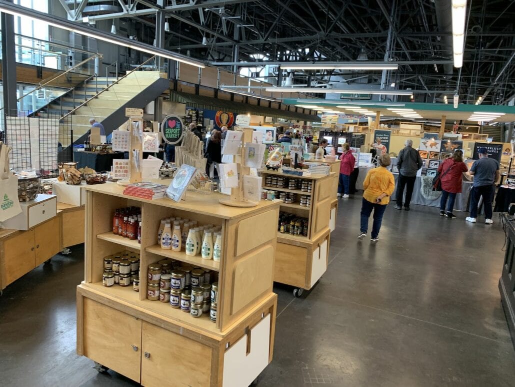 An indoor market near the bustling Halifax Cruise Port features various stalls and shelves with jars and bottles. Docked cruise passengers can explore as people browse goods beneath the industrial ceiling with visible beams and lights. A staircase leads to an upper level, adding to the adventure of what to do next.