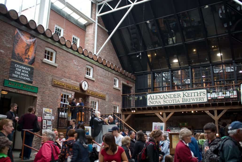 Amidst the bustling indoor space at Alexander Keith's Nova Scotia Brewery, near the Halifax Cruise Port, visitors gather to chat beneath brick walls. A large brewery sign and a clock adorn the doorway, while stairs and railings divide levels, making it an iconic stop for those docked in Halifax.