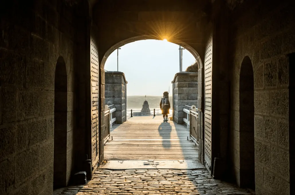 A person walks through a stone archway towards a sunlit boardwalk, where docked activities at Halifax Cruise Port await. The setting sun casts long shadows on the cobblestone path, and the sea is visible in the background.