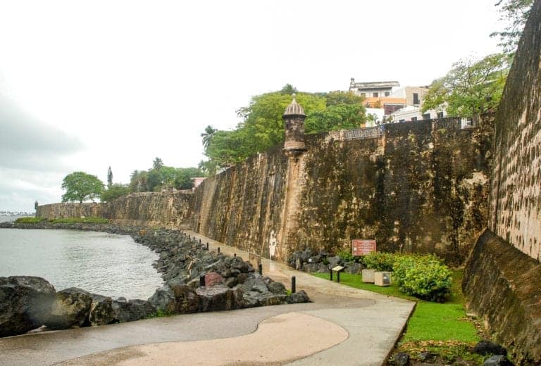 A stone fortress wall runs along a rocky shoreline under a cloudy San Juan sky. A walking path curves beside the wall, lined with greenery. A small turret juts out from the wall, while cruisers dock in the distance and buildings are visible in the background of Puerto Rico's vibrant scene.