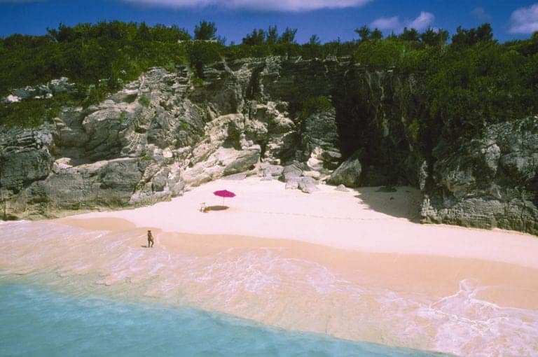 A sandy beach with turquoise water, a pink umbrella, and a lounge chair next to rocky cliffs and lush green vegetation brings a taste of the tropics. One person stands at the water’s edge, reminiscent of serene moments experienced on Bahamas cruises under a clear blue sky.