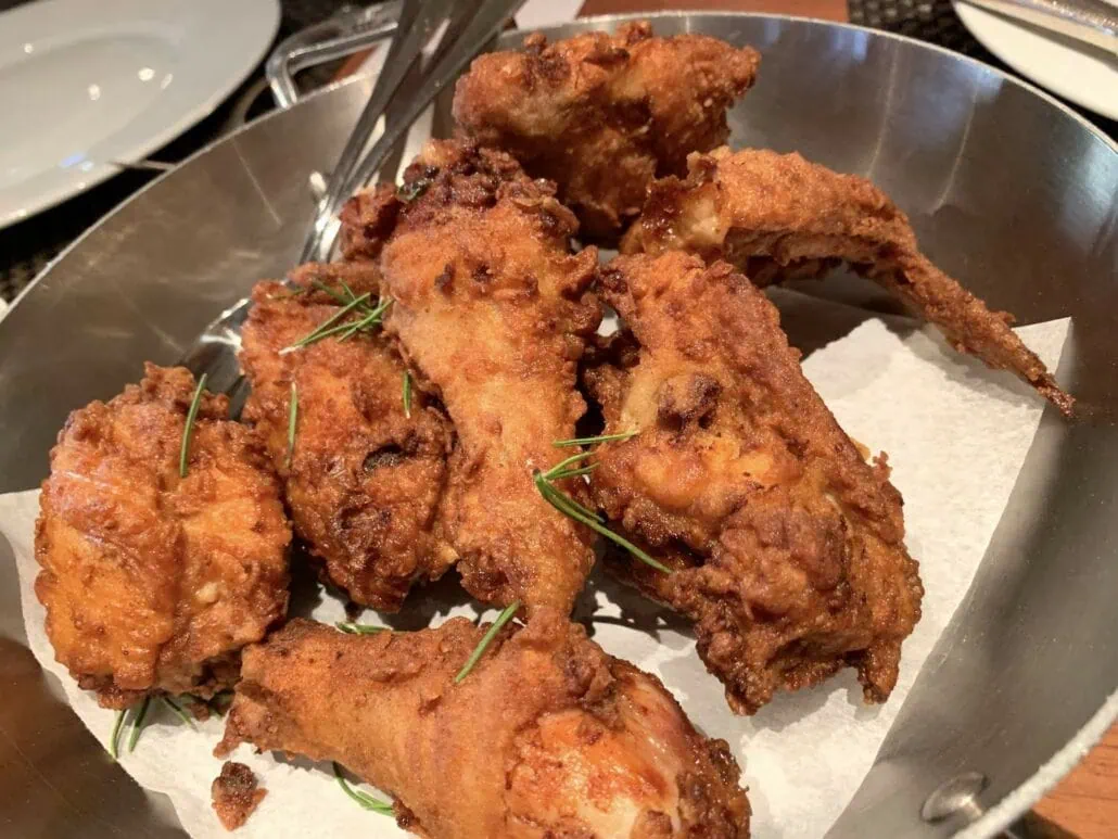 A stainless steel bowl filled with crispy fried chicken pieces, garnished with sprigs of rosemary and evoking the flavors you'd crave at the Haines Alaska Cruise Port, sits on a paper towel. A pair of metal tongs rests in the bowl, with a white plate visible in the background.
