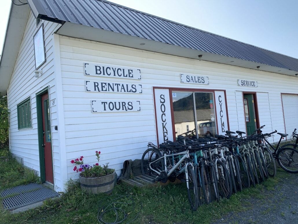 A white building near the Haines, Alaska cruise port offers bicycle rentals, sales, and tours. Several bicycles are parked in front of its metal roof, while a potted plant with pink flowers welcomes visitors at the entrance.