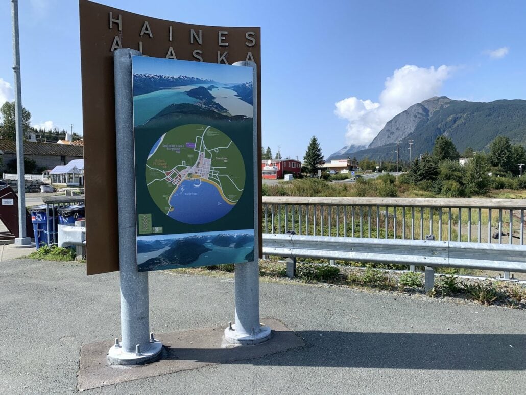 A metal display stand features a colorful map of Haines, Alaska, highlighting its charm as a serene cruise port. Behind the map, a large sign reads "Haines Alaska." The background showcases a mountainous landscape, quaint buildings, and a blue sky with drifting clouds.