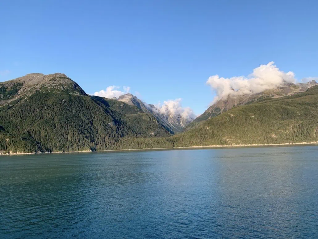 A serene landscape in Haines, Alaska, features a calm body of water in the foreground and a backdrop of lush, green mountains under a clear blue sky. Fluffy white clouds hover over the peaks near the cruise port, adding to the tranquil scene.