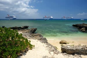A scenic view of a tropical beach in the Cayman Islands showcases clear turquoise waters, a rocky coastline, and lush greenery. Three large cruise ships float gently on the horizon under a partly cloudy blue sky.