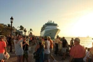 A large cruise ship is docked near a bustling waterfront in Key West. Numerous people are gathered on the shore, engaging in various activities, including taking photos and conversing. The sun is setting, casting a golden hue over the scene as palm trees line the shore, showcasing the vibrancy of the cruise industry.