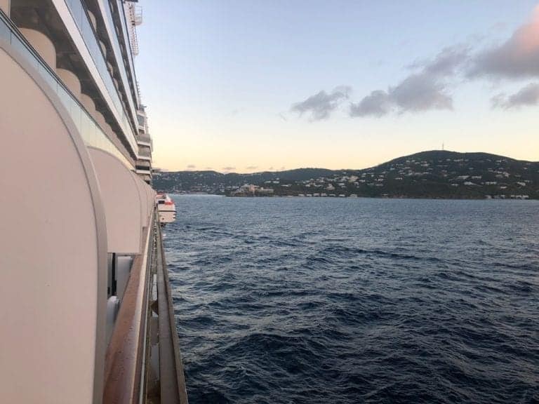 A view from the side deck of a cruise ship overlooking a calm sea with the distant coastal landscape of St. John. The sky is mostly clear with a few clouds visible, and the coast features rolling hills and scattered buildings, offering a picturesque scene typical of USVI charm.