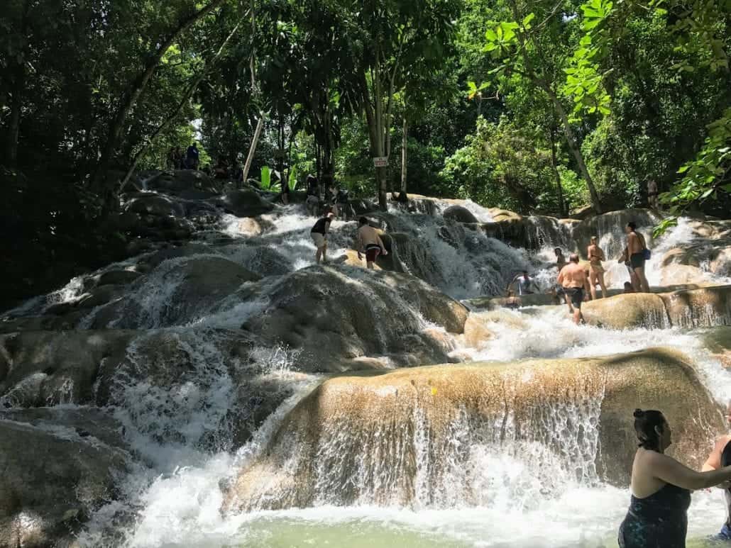 People are climbing and swimming at a multi-tiered waterfall surrounded by lush green vegetation. The cascades flow over smooth rocks, creating natural pools and streams. In the warmth of summer, some people are reaching the top while others are wading in the water below.