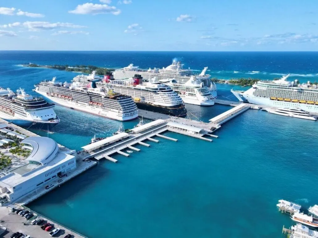 Aerial view of Nassau's bustling cruise ship port with multiple large ships docked. The clear blue ocean surrounds the vessels, and a few clouds are visible in the blue sky. White cruise ships and port buildings contrast beautifully with the turquoise water.