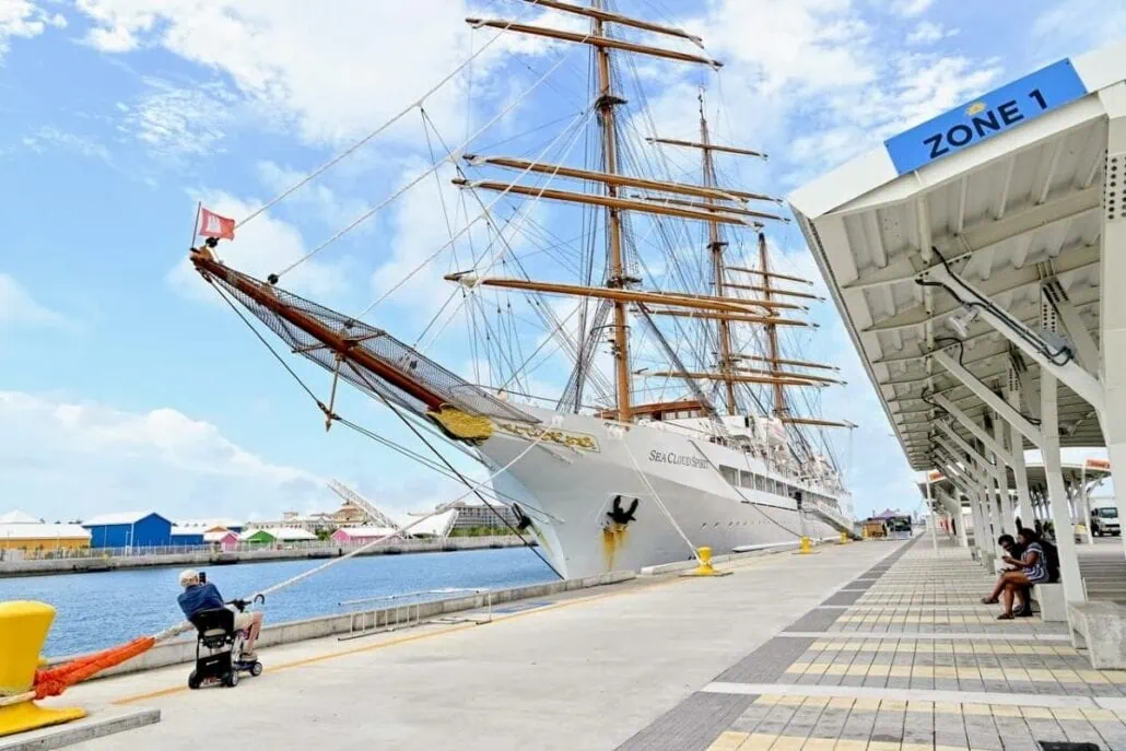 A large sailing ship is docked at Nassau's port next to a covered walkway labeled "Zone 1." A person in a mobility scooter gazes at the ship. In the background, colorful buildings stand against a clear blue sky.