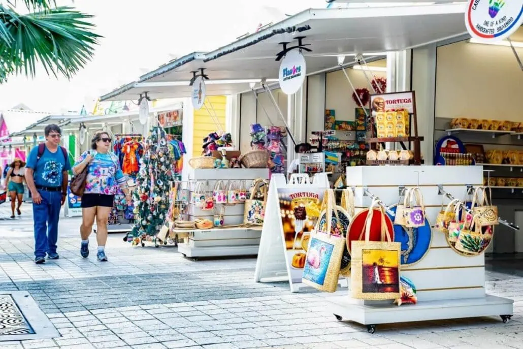 A couple strolls past outdoor market stalls in Nassau, offering colorful souvenirs and bags. The lively stalls display various goods, including crafts, snacks, and decorative items. A palm tree is visible in the top left corner, evoking a tropical ambiance typical of this vibrant locale.