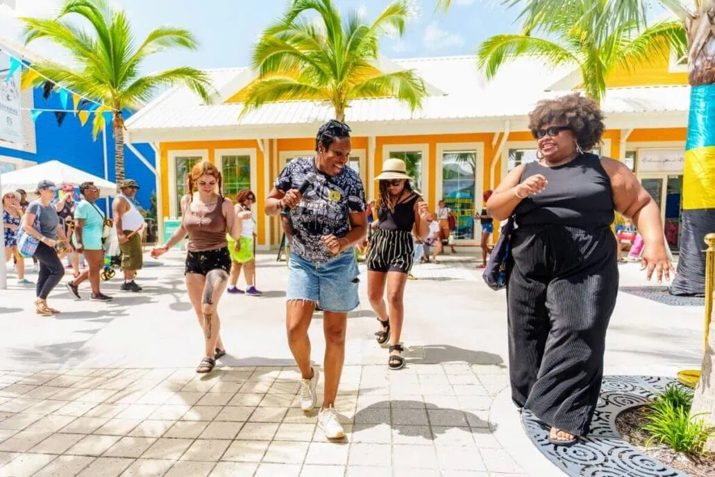 A group of people is dancing outdoors on a sunny Nassau day. They are in a courtyard with palm trees and colorful buildings in the background. The scene is lively, with others watching and enjoying the vibrant atmosphere.