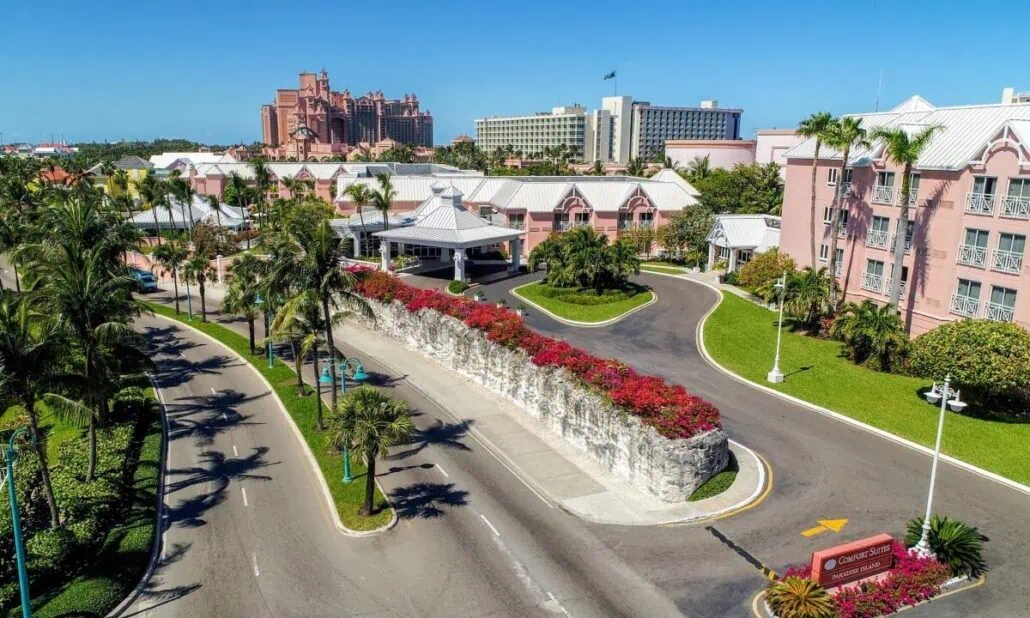 Aerial view of a Nassau resort featuring multiple pink buildings nestled among palm trees. A road with a central rock garden lined with red flowers winds through the area, while the sky remains clear and blue.