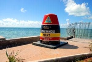 A large, colorful buoy-shaped monument marks the Southernmost Point of the continental USA in Key West, Florida. The text on the buoy indicates it's 90 miles to Cuba. With the ocean visible in the background and a partially cloudy sky above, this is one of the popular things to do in Key West.