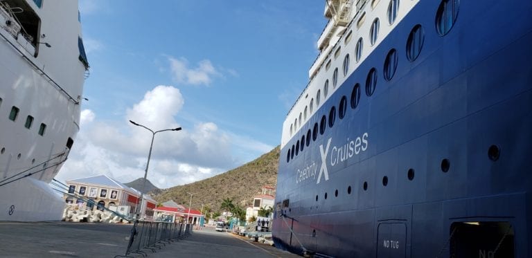 Two large cruise ships docked at a port by a hillside under a partly cloudy sky. To the left is a white cruise ship, and on the right is the navy-blue Celebrity Millennium with "Celebrity Cruises" printed on the side. A port building and streetlights are visible in the background, marking the start of an unforgettable trip.