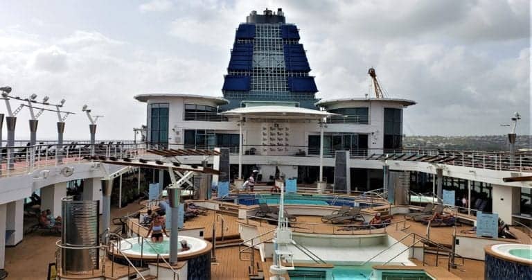 A deck of the Celebrity Millennium cruise ship features multiple pools, jacuzzis, and sunbathing areas illuminated by the sun and populated with a few guests. In the background, there is a large blue structure and some lounge chairs. The sky is partially cloudy, enhancing the relaxed atmosphere and entertainment options.