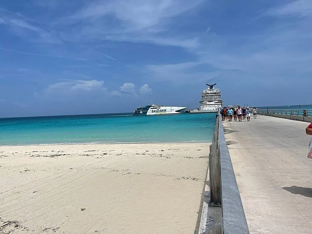 A sandy beach stretches into turquoise waters with a large cruise ship and a smaller boat near Bimini Cruise Port. People walk along a pier that extends from the beach towards the docked vessels under a blue sky with scattered clouds, offering everything you need to know for an unforgettable experience.