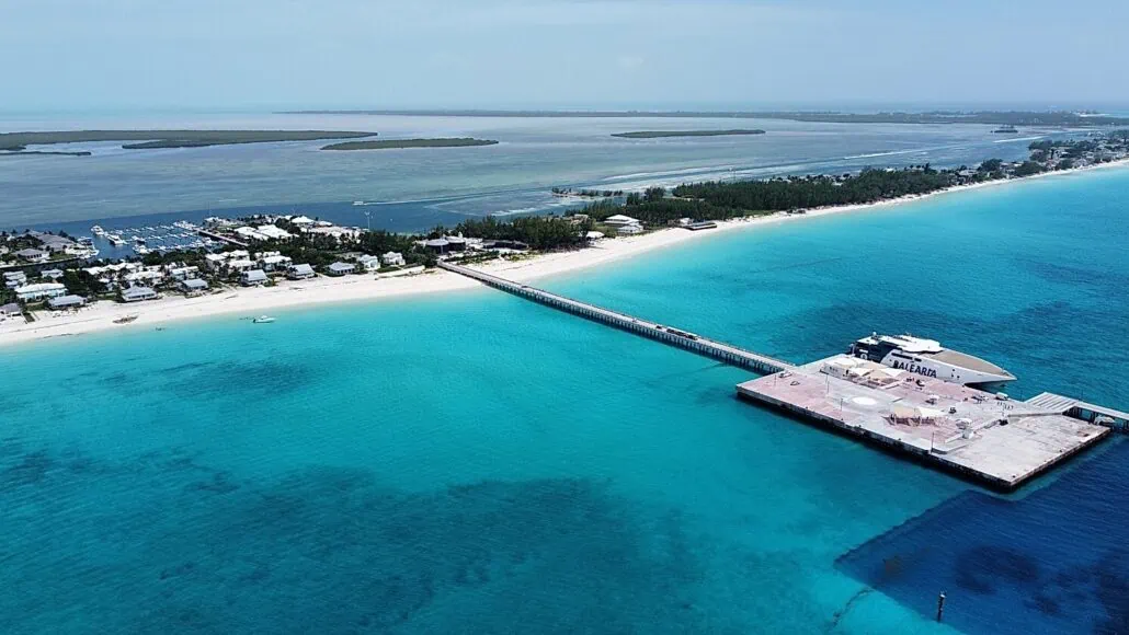 Aerial view of Bimini Cruise Port with clear turquoise water. A long pier extends from the sandy beach to a building on the water. Residential buildings and boats are visible on the left side, and an expanse of water and land is in the background.
