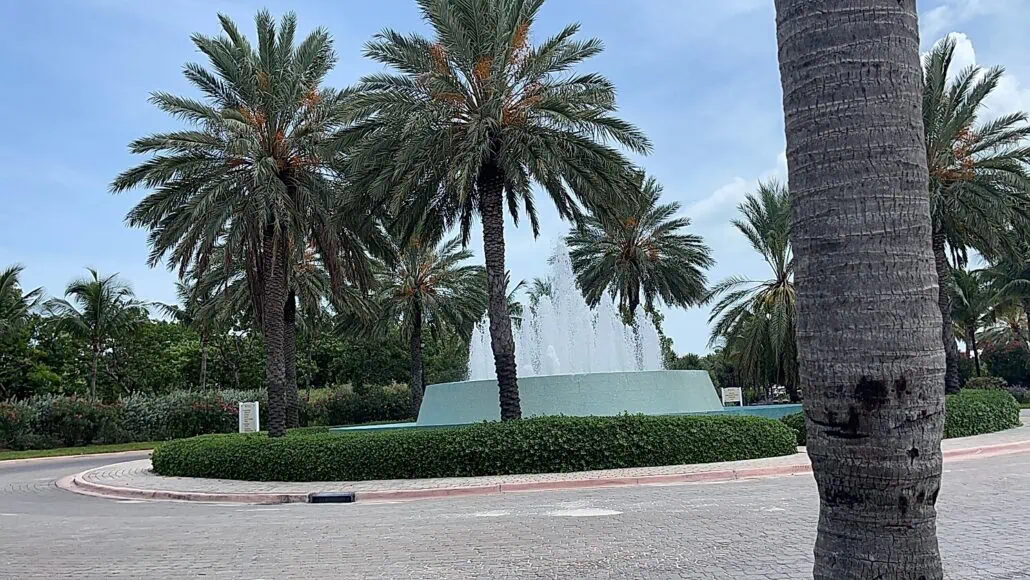 A circular water fountain surrounded by tall palm trees is situated on a brick-paved roundabout at the Bimini Cruise Port. In the background, more greenery is visible against a blue sky with some clouds. The scene appears peaceful and well-maintained.