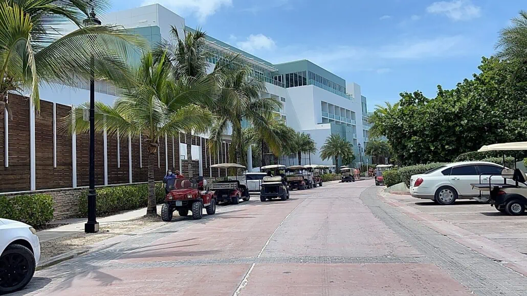A wide street lined with golf carts and a few cars under a sunny sky near the Bimini Cruise Port. The street has palm trees and modern white buildings on one side, with green trees on the other. People in bright clothing are seen among the parked golf carts, enjoying everything you need to know about this paradise.