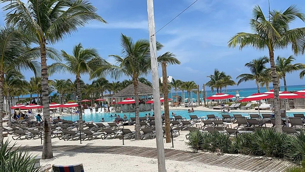 A beach resort with a large swimming pool is surrounded by palm trees, sun loungers, and red-and-white umbrellas. People are relaxing by the pool under a partly cloudy blue sky, enjoying everything you need to know for a perfect getaway. The ocean is visible in the background.