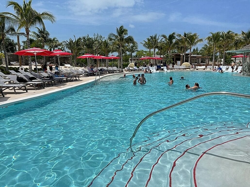 A clear swimming pool with people swimming and sitting by the edge under a sunny sky at the Bimini Cruise Port. The pool is surrounded by lounge chairs and red umbrellas. There are palm trees in the background, and beyond the pool area is a resort-like setting with more seating—everything you need to know for a perfect day.