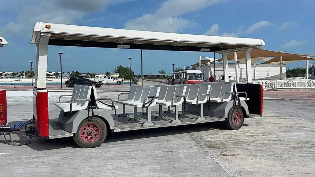 A red and white open-air tram with gray plastic seats is parked in an outdoor area. The tram, a popular feature of the Bimini Cruise Port, has a white roof and is positioned on a concrete surface with buildings, a fence, and blue sky in the background.