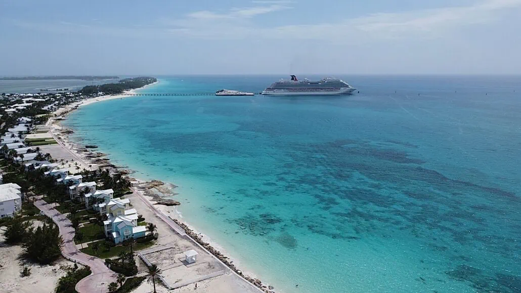 Aerial view of the Bimini Cruise Port, showcasing clear turquoise waters and white sandy beaches. A large cruise ship is docked near a long pier extending into the sea, with several white buildings with green roofs visible along the shore—a perfect travel destination.