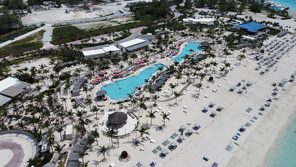 Aerial view of a resort with a large pool surrounded by red and white lounge chairs and umbrellas. Located near the Bimini Cruise Port, the resort is adjacent to a sandy beach lined with additional lounge chairs and umbrellas. The surrounding area features several buildings and lush greenery.