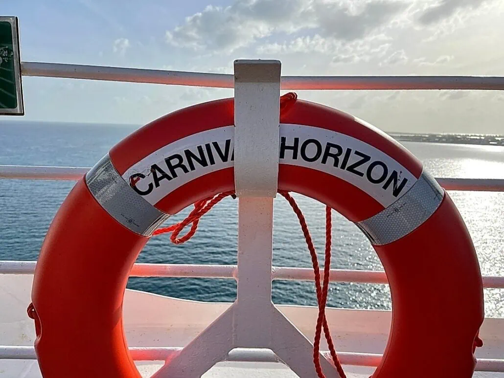 A red life buoy with the name "Carnival Horizon" is attached to a ship's railing. The bright sunlight illuminates the scene, and the calm ocean near Bimini Cruise Port is visible in the background along with the distant horizon.