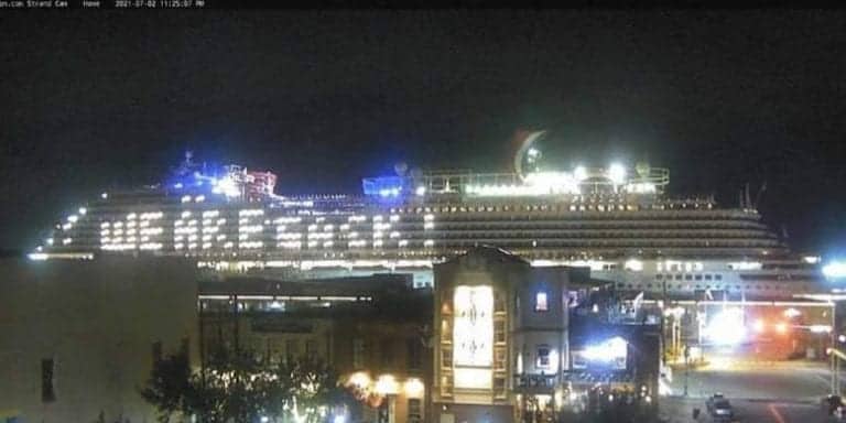 A large, brightly lit cruise ship docked at Galveston port at night. The Carnival Vista's lights illuminate the surroundings, including nearby buildings. The sky is dark, and the overall scene appears serene and calm with the lights creating a lively atmosphere.