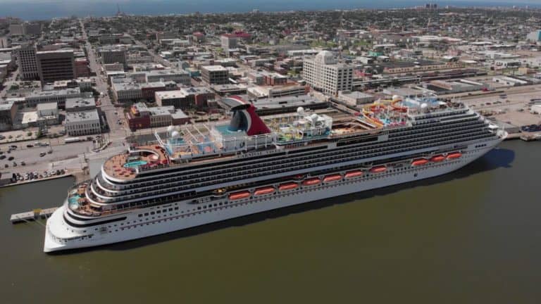 A large Carnival Cruise Line ship is docked at a bustling port in an urban area, its multiple decks, colorful slides, and distinctive red and black funnel are ready for a grand celebration. Surrounding the port are various buildings and infrastructure, with the sea visible in the distance.