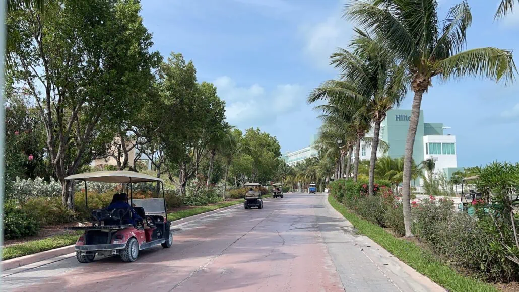 A paved road lined with palm trees and greenery, with several golf carts driving on it, leads to a light-colored building with "Hilton" visible on the facade. The setting near Bimini Cruise Port showcases clear skies dotted with a few clouds.