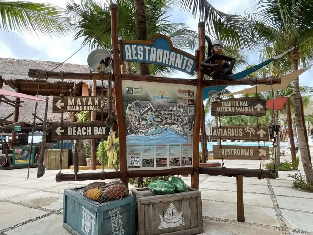 A wooden signpost at the Costa Maya resort displays directions to restaurants, a Mayan healing rituals area, beach bay, a traditional Mexican market, aviarius, and restrooms. The lush setting resembles something from a Celebrity Edge trip report with tropical plants and thatched structures in the background.