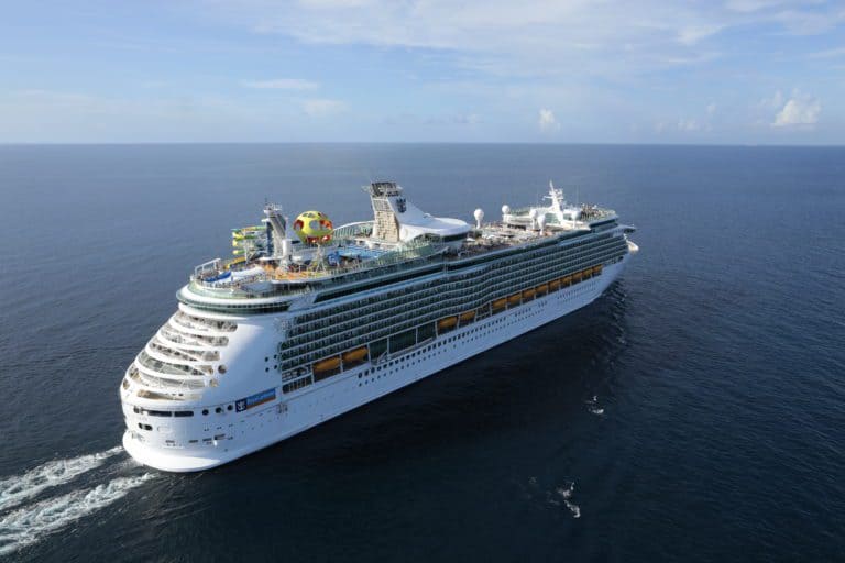 Aerial view of a large white Royal Caribbean cruise ship sailing in the open ocean. The ship features multiple decks with various amenities, including a waterslide and recreational facilities on the top deck. The sky is clear, and the water is calm.