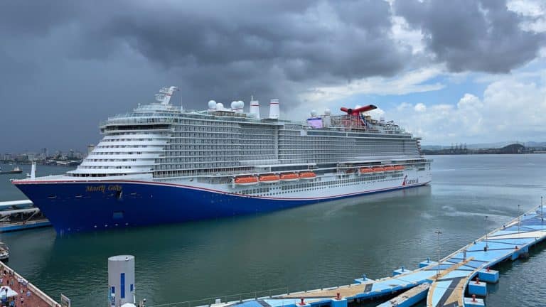 A large cruise ship named "Mardi Gras" is docked at Puerto Rico's port under a cloudy sky. With multiple decks, visible lifeboats, and its distinctive red and blue color scheme, the scene offers a picturesque view of the port and surrounding water.