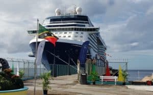 A large cruise ship, the first to arrive in 18 months, is docked at a port adorned with various flags, including one from Saint Kitts and Nevis. Nearby, several potted plants and a bench grace the water's edge under a partly cloudy Caribbean sky.