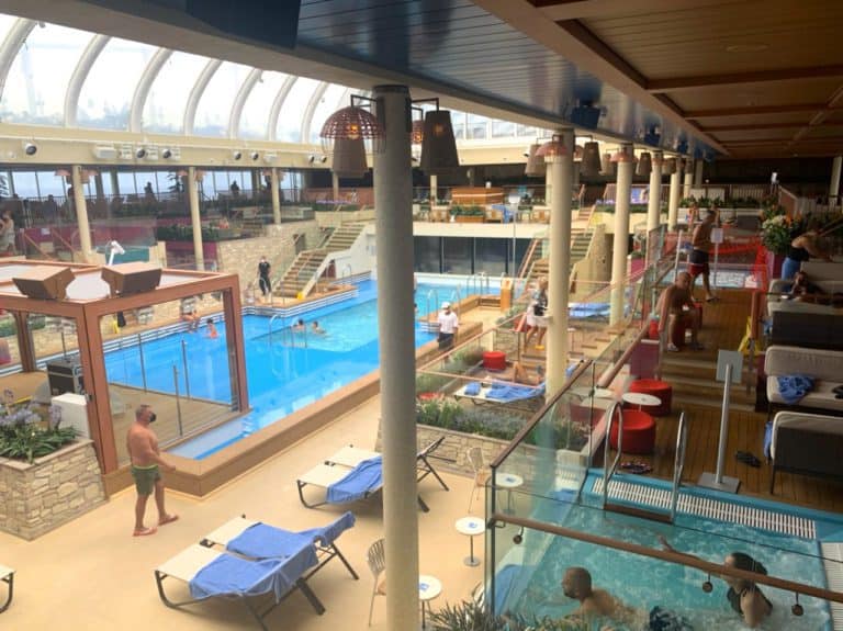 A spacious indoor pool area with clear blue water, surrounded by lounge chairs, tables, and plants. Several people are relaxing, swimming, and walking around. There is a glass roof allowing natural light in and a balcony overlooking the pool. It feels like you're cruising Italian style on the Costa Smeralda.