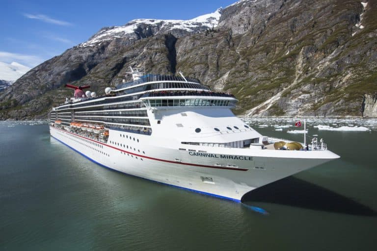 A large cruise ship named Carnival Miracle is sailing through the calm waters of Alaska, surrounded by steep, rocky mountains with patches of snow. The ship is white with a red and black funnel and a red stripe along its side. The sky is clear and blue.
