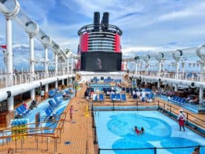 The deck of the Disney Fantasy buzzes with activity: passengers relish in the pool and lounge chairs while a large movie screen entertains them. The ship's funnel stands tall under a partly cloudy sky, creating the perfect backdrop for an unforgettable day at sea.