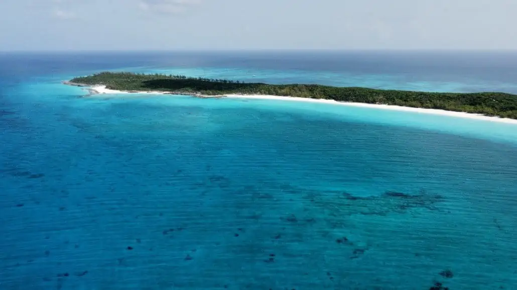 Aerial view of Half Moon Cay, a long, narrow island surrounded by turquoise water. The island features a dense green forest area, white sandy beaches, and clear blue ocean extending into the horizon. The sky is mostly clear with a few clouds—perfect for your Carnival vacation photos.