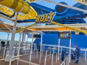 An amusement park ride entrance features a sign that reads "BOLT Ultimate Sea Coaster." The area includes a waiting line with metal railings, a digital display with ride information, and an attendant in a blue uniform. The bright yellow roller coaster track is visible above, reminiscent of one you'd find on a cruise ship.