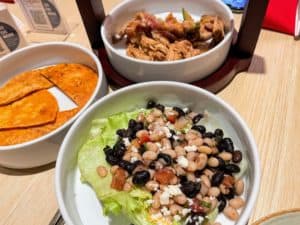 A close-up of a Chibang! table with three dishes. The foreground shows a bowl of mixed beans on a bed of lettuce, topped with cheese. To the left is a plate of sliced, seasoned potatoes. In the background is a bowl of shredded meat—perfect for your next Restaurant Review!