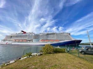 A large Carnival Mardi Gras cruise ship is docked next to a grassy area with a few small trees and rocks. There are people and cars nearby. The sky above is blue with scattered clouds.